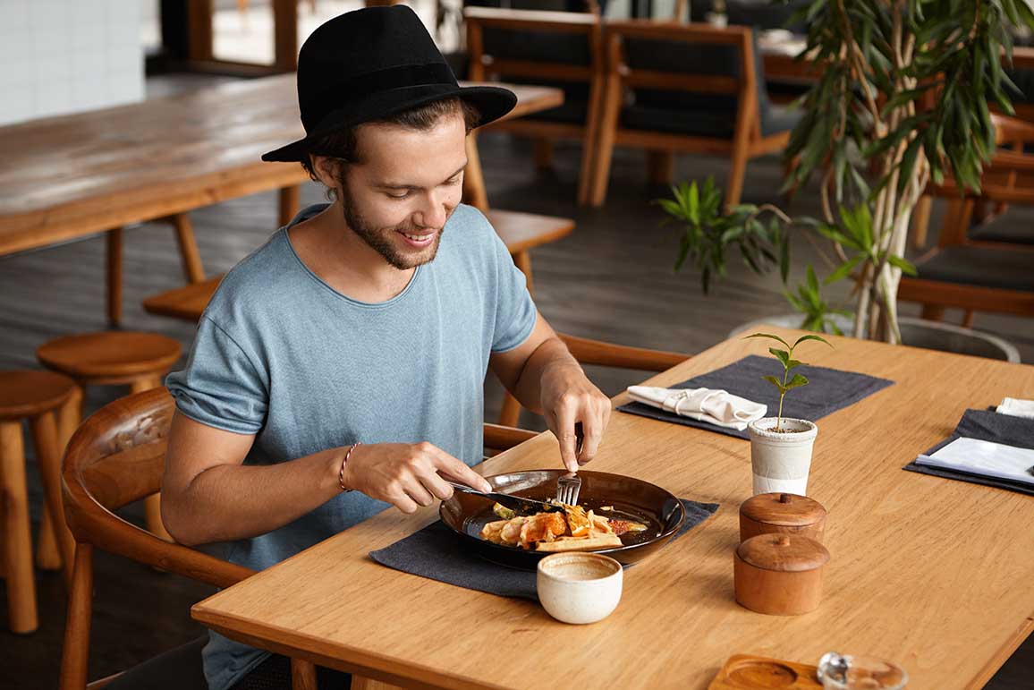Indoor shot of happy young student wearing stylish hat eating delicious food with knife and fork during break at university canteen, enjoying fresh and healthy lunch, sitting at wooden table alone